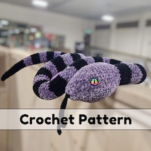 May include: Crochet pattern for a purple and black striped snake amigurumi. The snake has a rainbow eye and is curled up in a coil.