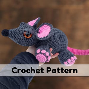 May include: Crocheted gray mouse with pink ears, paws, and tail. The mouse has large orange eyes. The image includes the text "Crochet Pattern".