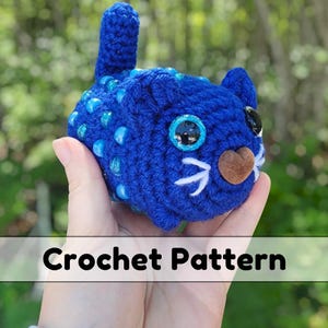 May include: A handmade blue crochet cat with blue bead embellishments. The cat features large, round eyes, a brown nose, and white whiskers. The words "Crochet Pattern" are displayed at the bottom of the image.