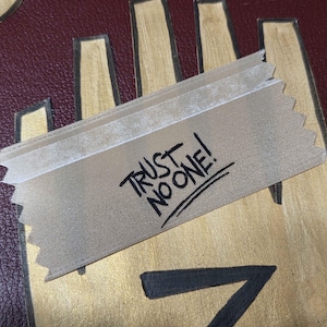 May include: A tan ribbon with the words "TRUST NO ONE!" in black handwritten text. The ribbon is placed over a gold and burgundy background with a hand-like design.