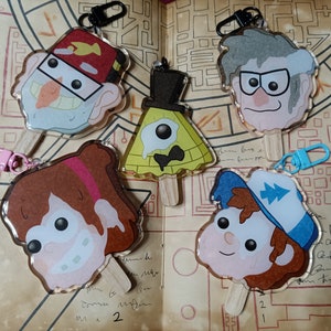 May include: Five colorful character keychains from the show Gravity Falls. The characters are: Stan Pines, Bill Cipher, Mabel Pines, Dipper Pines, and Ford Pines. Each keychain is a clear plastic circle with a character image and a wooden stick.