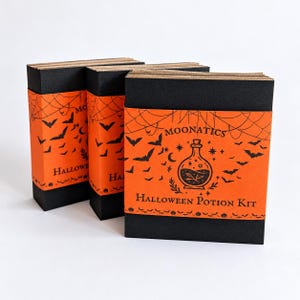 May include: Three rectangular boxes with black covers and orange paper bands. The bands feature black illustrations of bats, spiderwebs, and a potion bottle with the text MOONATICS HALLOWEEN POTION KIT printed in black ink.
