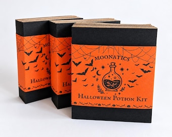 Pode incluir: Três caixas retangulares com tampas pretas e faixas de papel laranja. As faixas apresentam ilustrações pretas de morcegos, teias de aranha e um frasco de poção com o texto MOONATICS HALLOWEEN POTION KIT impresso em tinta preta.