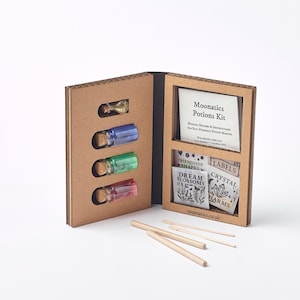 Può includere: Un kit di pozioni Moonatics, presentato in una scatola di cartone. Contiene quattro piccole bottiglie di vetro con contenuto colorato e tappi di sughero. Il kit include istruzioni, etichette e bastoncini di legno. Il testo "Moonatics Potions Kit" è visibile.