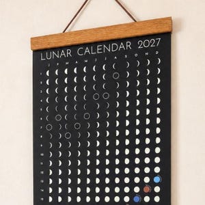 May include: A black wall calendar titled LUNAR CALENDAR 2027 with white moon phase icons arranged in a grid. The calendar is held by a wooden magnetic hanger at the top with a brown leather cord for hanging. It displays months, days, and phases.