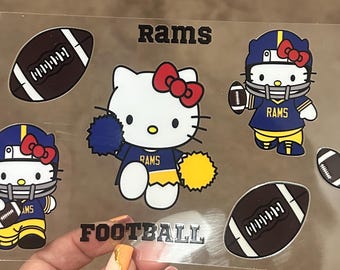 Può includere: Un set di otto adesivi con Hello Kitty vestita da giocatrice di football. Gli adesivi sono blu, gialli e bianchi con le parole "Rams" e "Football".
