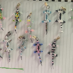 May include: A collection of beaded lizard charms in various colors, including green, blue, pink, and black. Each charm has a gold-colored clasp for attachment. The lizards are crafted with small beads, creating a textured appearance.
