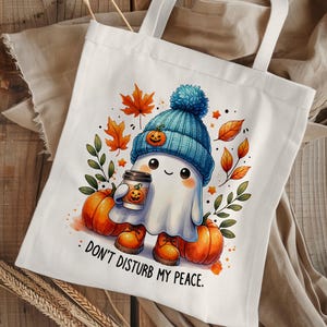 Op de afbeelding: Witte tote bag met een schattig spookje dat een blauw-witte hoed draagt en een kopje koffie vasthoudt. Het spookje is omringd door herfstbladeren en pompoenen. De tekst op de tas luidt "Stoor me niet."