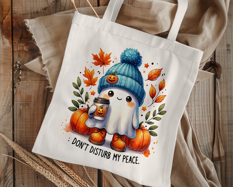 Op de afbeelding: Witte tote bag met een schattig spookje dat een blauw-witte hoed draagt en een kopje koffie vasthoudt. Het spookje is omringd door herfstbladeren en pompoenen. De tekst op de tas luidt "Stoor me niet."