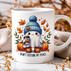 Op de afbeelding: Een witte keramische mok met een schattig cartoon-spookje dat een blauwe hoed draagt en een kopje koffie vasthoudt. Het spookje is omringd door herfstbladeren en pompoenen. Op de mok staat "Stoor me niet."