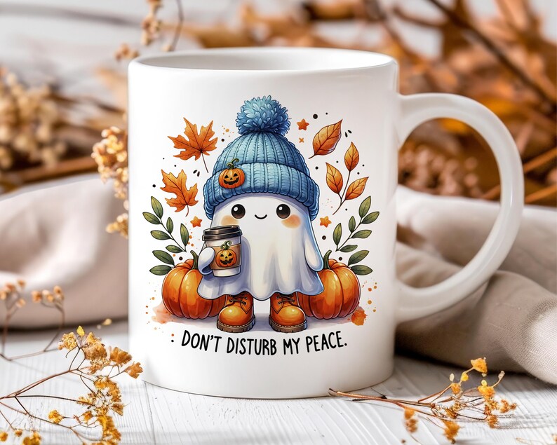 Op de afbeelding: Een witte keramische mok met een schattig cartoon-spookje dat een blauwe hoed draagt en een kopje koffie vasthoudt. Het spookje is omringd door herfstbladeren en pompoenen. Op de mok staat "Stoor me niet."