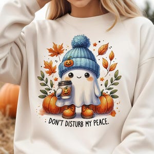 Op de afbeelding: Wit sweatshirt met een schattig spookje dat een blauwe muts draagt en een kopje koffie vasthoudt. Het spookje is omringd door herfstbladeren en pompoenen. De tekst "Stoor mijn rust niet" staat onder het spookje.