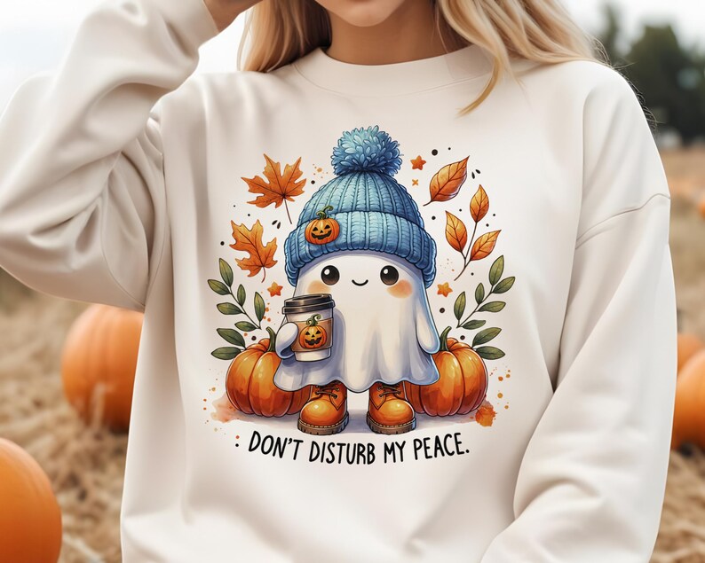 Op de afbeelding: Wit sweatshirt met een schattig spookje dat een blauwe muts draagt en een kopje koffie vasthoudt. Het spookje is omringd door herfstbladeren en pompoenen. De tekst "Stoor mijn rust niet" staat onder het spookje.