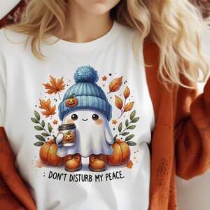 Op de afbeelding: Wit T-shirt met een schattig cartoon-spookje dat een blauw-witte hoed draagt en een pumpkin spice latte vasthoudt. Het spookje is omringd door herfstbladeren en pompoenen. De tekst "Stoor mijn rust niet" is onder het spookje gedrukt.