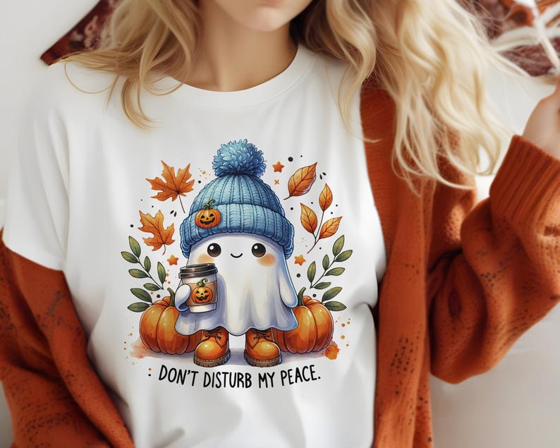 Op de afbeelding: Wit T-shirt met een schattig cartoon-spookje dat een blauw-witte hoed draagt en een pumpkin spice latte vasthoudt. Het spookje is omringd door herfstbladeren en pompoenen. De tekst "Stoor mijn rust niet" is onder het spookje gedrukt.