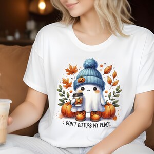 Op de afbeelding: Wit T-shirt met een schattig spookje dat een blauwe muts draagt en een pumpkin spice latte vasthoudt. Het spookje is omringd door herfstbladeren en pompoenen. De tekst op het T-shirt luidt "Stoor mijn rust niet."