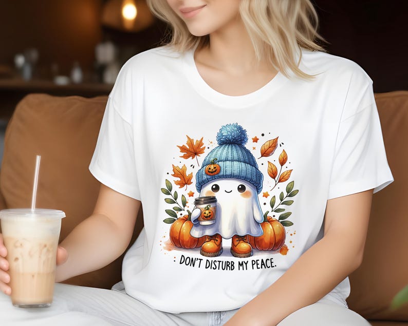 Op de afbeelding: Wit T-shirt met een schattig spookje dat een blauwe muts draagt en een pumpkin spice latte vasthoudt. Het spookje is omringd door herfstbladeren en pompoenen. De tekst op het T-shirt luidt "Stoor mijn rust niet."