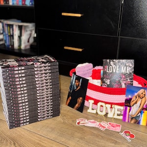 May include: A stack of books titled "LOVE ME NOT" next to a red and pink woven basket filled with items. The basket contains a photo, a card that says "LOVE ME", and stickers. The word "LOVE" is spelled out in wooden letters.