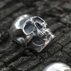 Könnte beinhalten: Drei kleine, metallische, totenkopfförmige Perlen oder Anhänger mit polierter Oberfläche. Jeder Totenkopf weist detaillierte Augenhöhlen, eine Nasenhöhle und Zähne auf, wobei die reflektierende Oberfläche die Konturen der Schädelform betont.