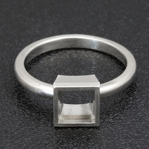 Puede incluir: Un anillo de plata con un engaste cuadrado. La banda del anillo tiene un diseño simple y redondeado. El engaste cuadrado es hueco, creando una forma geométrica abierta. El anillo se muestra sobre un fondo oscuro.