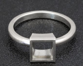 May include: A silver ring with a square setting. The ring band is a simple, rounded design. The square setting is hollow, creating an open, geometric shape. The ring is displayed against a dark background.