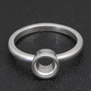 Puede incluir: Un anillo de plata con un engaste circular. El anillo tiene una banda simple y un engaste circular abierto prominente, diseñado para sostener una piedra preciosa u otro elemento decorativo. El anillo se muestra sobre un fondo oscuro.