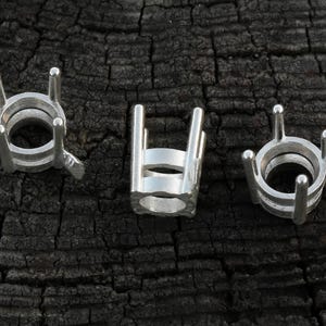 May include: Three silver jewellery settings are shown against a dark, textured background. Each setting has a circular base with four prongs extending upwards, designed to hold a gemstone. The settings are made of a shiny metal.