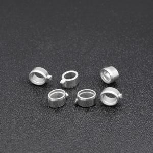 May include: Six small, silver-coloured metal rings with a circular design are scattered on a dark surface. Each ring has a small, protruding tab. The rings appear to be new and unused.