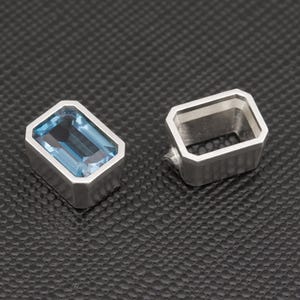 Puede incluir: Dos anillos plateados de forma rectangular. Un anillo presenta una piedra preciosa rectangular de color azul claro. El otro anillo es un marco plateado vacío. Los anillos están sobre un fondo texturizado gris oscuro.