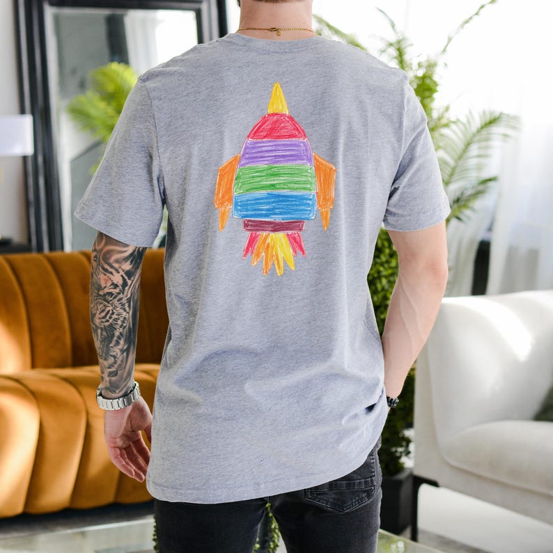 May include: A gray t-shirt featuring a colorful, hand-drawn rocket graphic on the back. The rocket design includes red, orange, yellow, green, blue, and purple elements. The shirt is short-sleeved and made of a soft material.