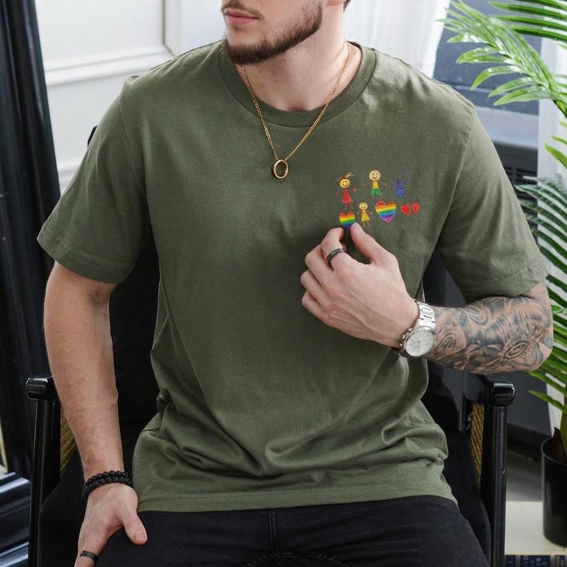 May include: Olive green t-shirt with a small embroidered design of stick figures, a rainbow, and hearts. The shirt has short sleeves and a crew neck. A person is wearing the shirt, and is also wearing a gold necklace and a watch.