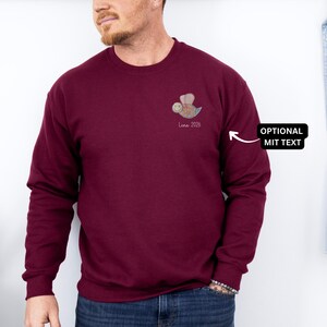 May include: Burgundy crewneck sweatshirt with a small bee graphic and the text "Luna 2026" on the chest. The bee is in shades of brown, blue, and yellow. The sweatshirt is a solid color and has long sleeves.
