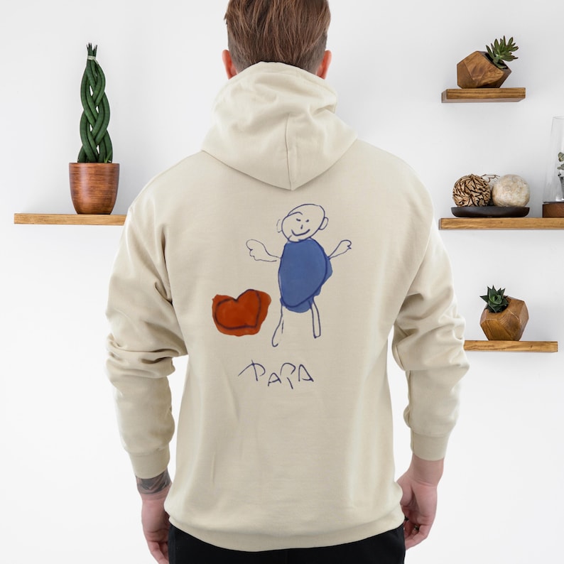 May include: Beige hoodie with a drawing on the back. The drawing features a blue figure, a red heart, and the word "PAPA" in black. The hoodie has a hood and long sleeves. The person is facing away from the camera.