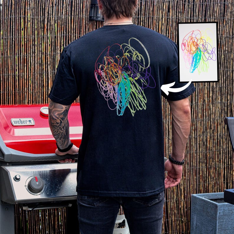 May include: Black short-sleeved t-shirt with a colorful abstract scribble design on the back. The design features a mix of blue, green, yellow, orange, and purple lines. A framed drawing with a similar design is also visible.