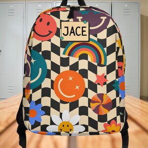 Op de afbeelding: Een kleurrijke rugzak met een zwart-wit geblokt patroon. De rugzak heeft smiley gezichten, regenbogen, sterren en bloemen. Een naamlabel leest "JACE".