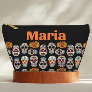 May include: A black zippered pouch featuring the name Maria in orange text at the top. The surface is decorated with a repeating pattern of colorful sugar skulls in shades of yellow, red, blue, and white, each with intricate floral and cross details.