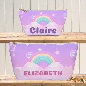 Könnte beinhalten: Zwei Kosmetiktaschen mit einem pastellfarbenen Regenbogen- und Wolkenmuster. Die obere Tasche trägt den Namen "Claire" in Lila, und die untere Tasche trägt den Namen "Elizabeth". Beide Taschen haben einen Reißverschluss und einen hellvioletten Hintergrund mit Sternen und Blasen.