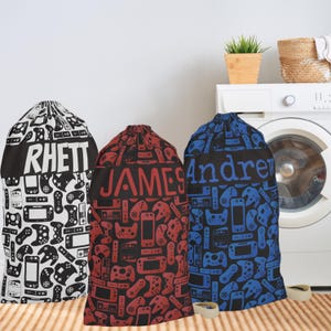 May include: Three drawstring laundry bags featuring a repeating pattern of video game controllers and handheld consoles. Each bag has a solid horizontal band displaying the names RHETT, JAMES, and Andrew in a bold, sans-serif font.