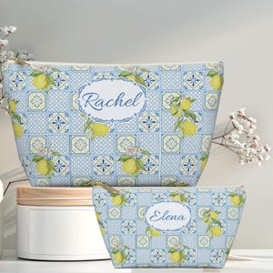 Op de afbeelding: Twee make-uptassen met ritssluiting met een blauw-wit tegelpatroon en citroenillustraties. De grotere tas heeft de naam "Rachel" in een witte ovaal, en de kleinere tas heeft de naam "Elena". Beide tassen hebben gouden ritsen.