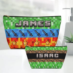 May include: Two pixelated cosmetic bags. The larger bag has the name "JAMES" in pixelated letters, with a green, blue, orange, and yellow design featuring swords and stars. The smaller bag has the name "ISAAC" in pixelated letters, with a green, brown, and black design.