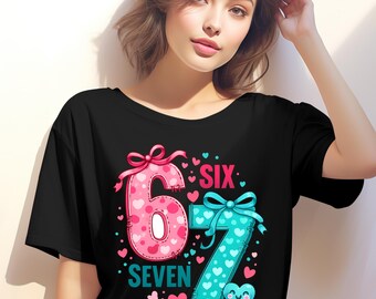 Peut inclure: T-shirt noir avec un motif graphique coloré représentant les chiffres "6" et "7" avec des cœurs. Le mot "SIX" est au-dessus du chiffre 7, et le mot "SEVEN" est en dessous du chiffre 6. Les chiffres sont décorés de nœuds.