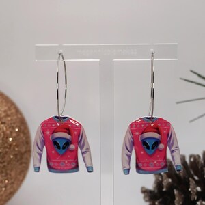 Könnte beinhalten: Ein Paar Creolen mit einem rosa Sweater-Design, das einen Alien mit Weihnachtsmütze zeigt. Die Ohrringe haben ein festliches Urlaubs-Thema, mit einem hellblauen Alien-Gesicht und weißen und hellblauen Akzenten. Die Ohrringe werden auf einem transparenten Ständer präsentiert.