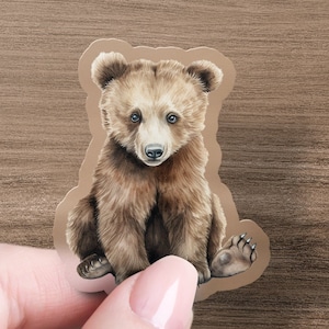 May include: A brown bear cub sticker with a white patch on its chest. The bear cub is sitting with its paws tucked in front of it and looking directly at the camera.