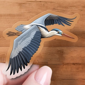 May include: A sticker of a grey heron in flight with its wings spread wide. The heron has a long, thin beak and a blue head. The sticker is on a wooden background.
