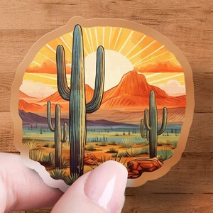 May include: A round sticker with a desert sunset scene featuring three tall cacti, a mountain range, and a sun with rays. The background is a gradient of orange, yellow, and blue.