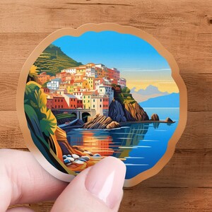 May include: A round sticker featuring a colorful illustration of a coastal Italian village. The artwork depicts buildings in shades of orange, yellow, and white, set against a blue sky and sea. The sticker has a light brown border and is held by a hand.