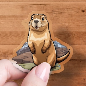 May include: A cartoon sticker of a prairie dog standing on its hind legs with a happy expression. The prairie dog is brown and white and is surrounded by rocks and grass.