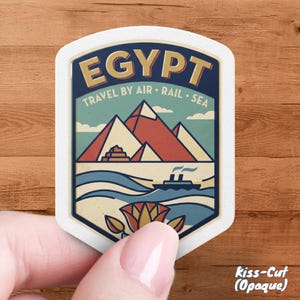 May include: A white, kiss-cut sticker with a vintage travel design. The sticker features the word "EGYPT" in gold lettering, pyramids, a boat, and the text "TRAVEL BY AIR • RAIL • SEA".