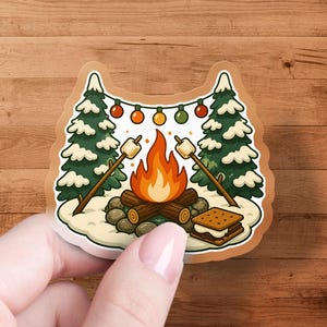 May include: A die-cut sticker featuring a campfire scene with two marshmallows on sticks, a s'more, and string lights hanging between two snow-covered pine trees. The illustration uses a color palette of orange, brown, green, and white.