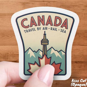 May include: A white, kiss-cut sticker with a vintage travel design. The sticker features the word "CANADA" in red, with the text "TRAVEL BY AIR • RAIL • SEA" below. The design includes a maple leaf, mountains, and a tower.